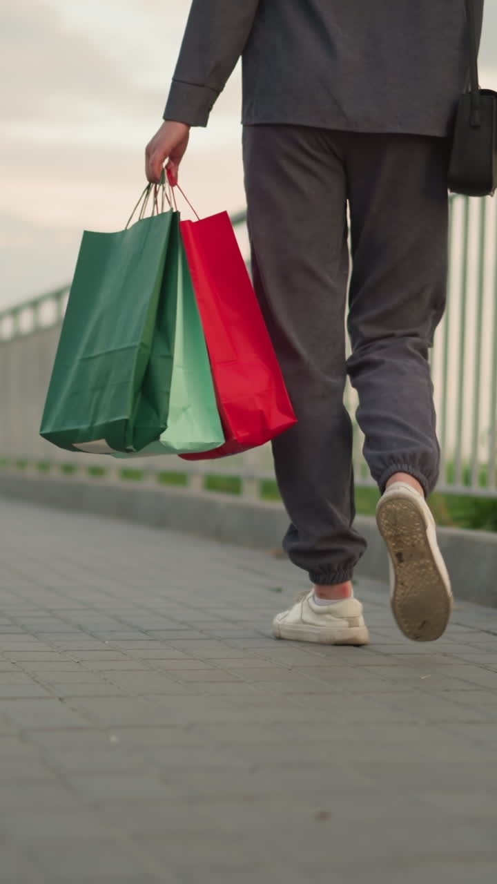 vista trasera de la pierna de la dama con ropa de color gris caminando a lo largo del carril de hierro con bolsas de compras en la mano y bolso de mano negro, creando una escena al aire libre casual con fondo borroso con vegetación distante