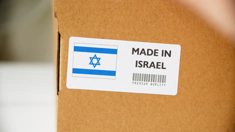 manos aplicando etiqueta de bandera hecha en israel en una caja de cartón de envío con productos