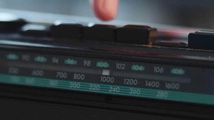 Primo piano a fuoco selettivo di un pannello sintonizzatore radio nero con numeri di frequenza luminosi, marcature del cursore e una superficie lucida riflettente sotto una morbida luce ambientale interna.
