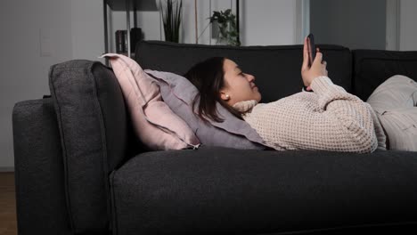 cute asian woman drops on the sofa and starts browsing on her smartphone