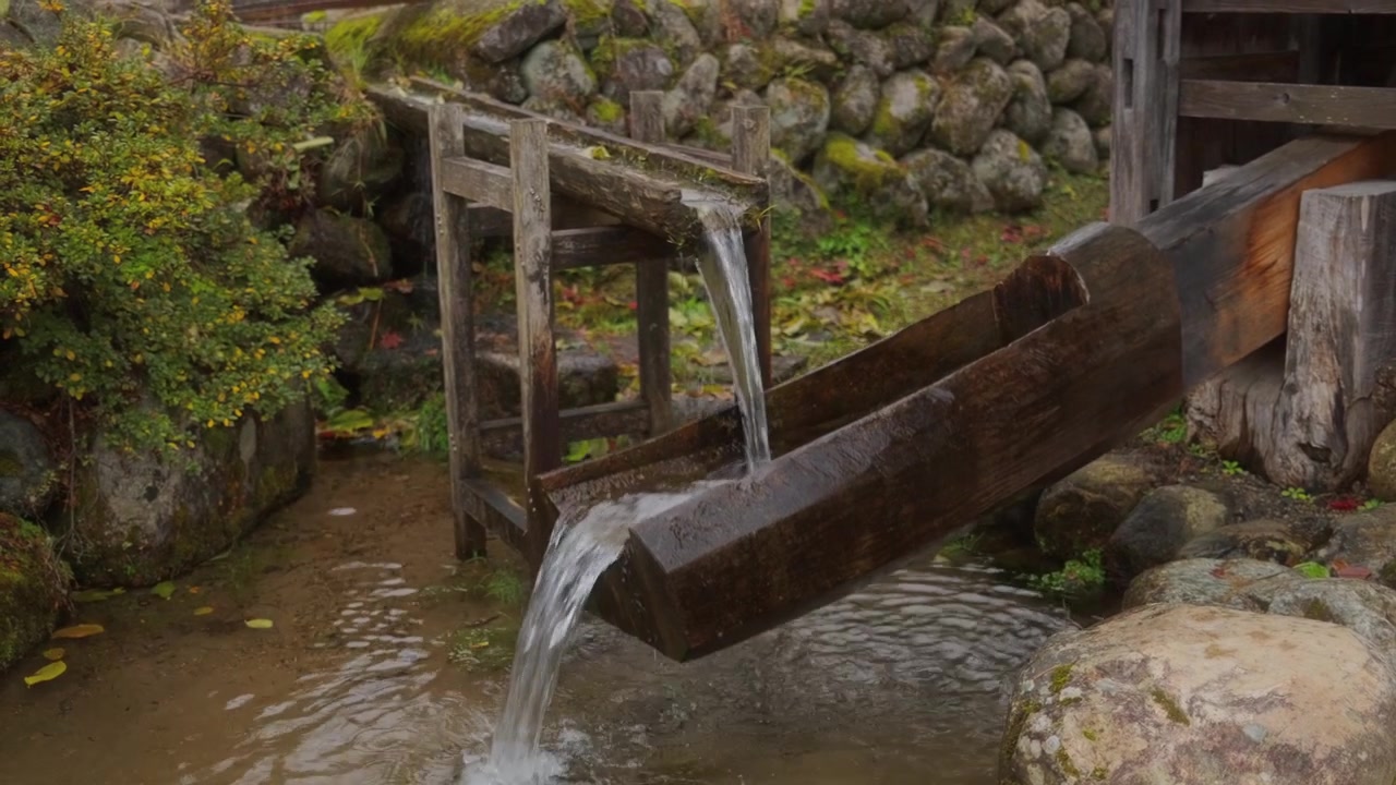 Vecchio canale d'acqua in legno con acqua fresca che scorre in un contesto di villaggio tradizionale giapponese. Dettaglio rustico di campagna con pietre, movimento del ruscello e un'autentica atmosfera storica.