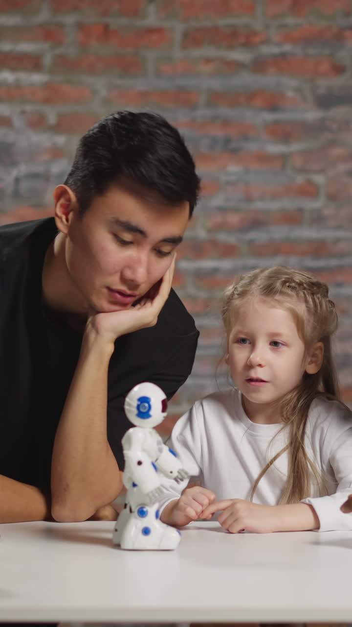 Blonde little girl with young Asian and African-American home teachers looks at funny toy robot on table with laptop computer in apartment
