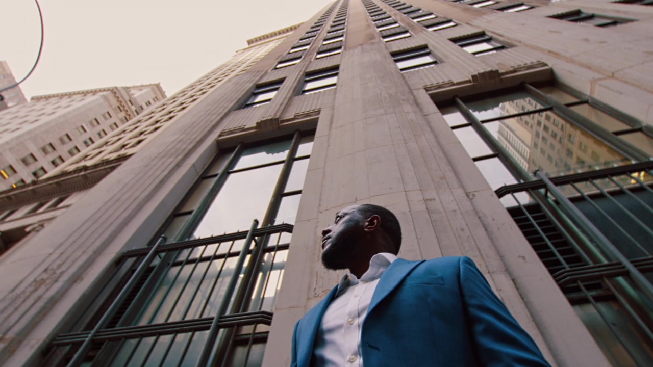 New York, USA, 1 May 2024: Businessman gazing upward beside art deco facade, New York. Low angle with green metallic railings and concrete columns converging overhead.