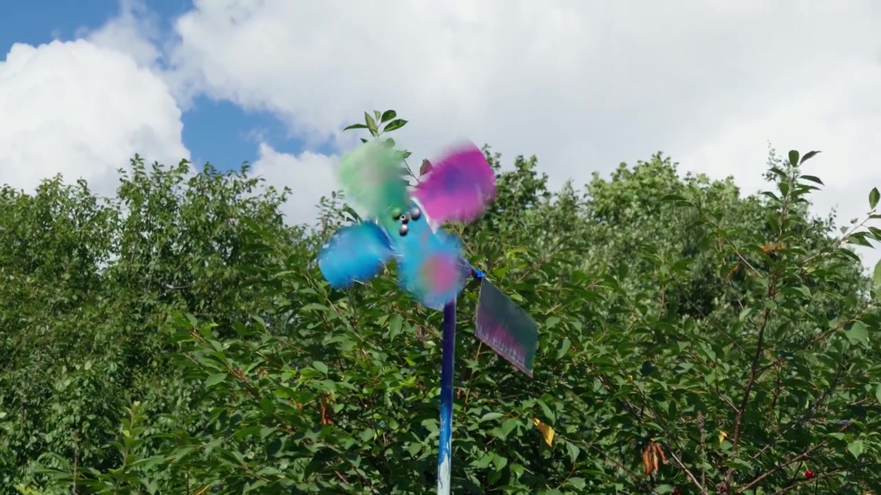 Girandola di plastica dai colori vivaci che gira rapidamente nella brezza estiva contro alberi verdi e rigogliosi e un cielo blu pieno di nuvole bianche, inquadratura media in campagna