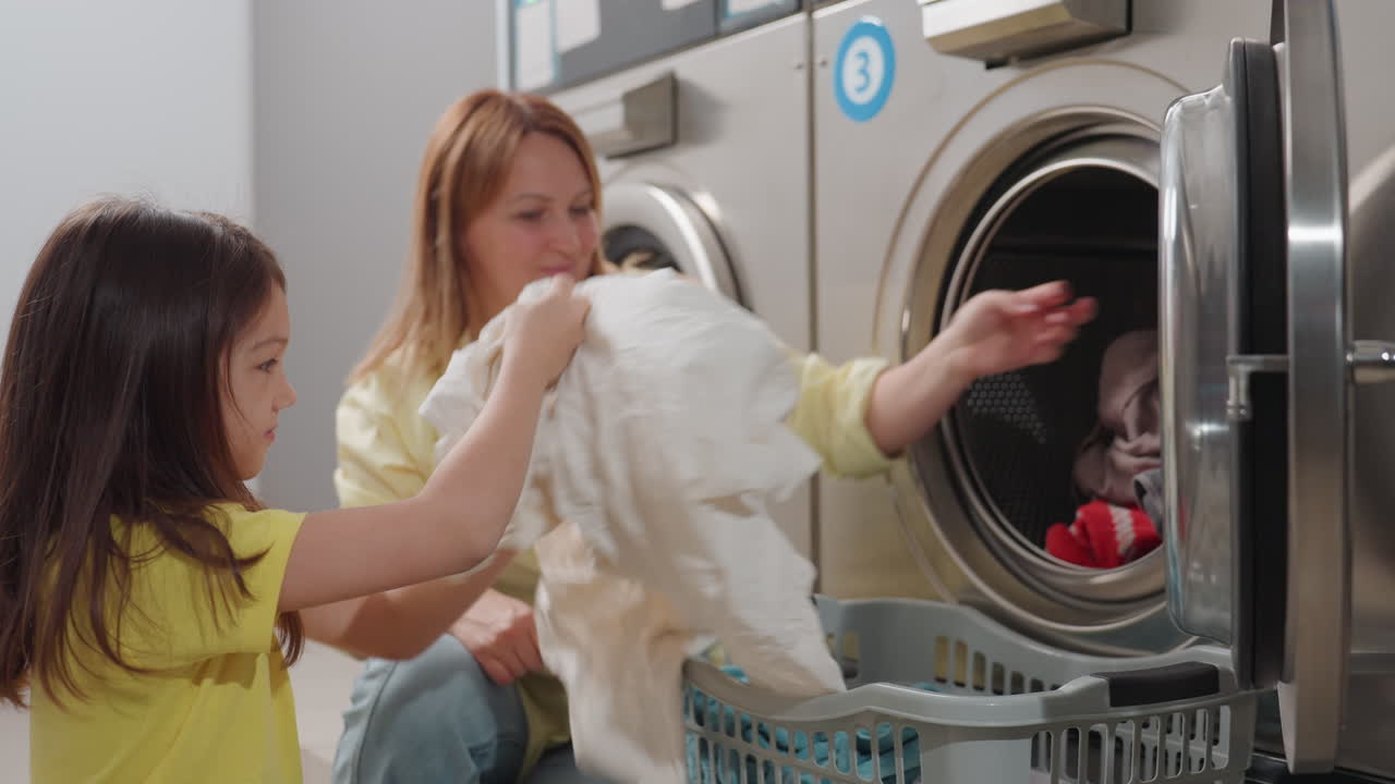Caregiver sits by washer guiding toddler as they load clothes, gray sheet swings from basket toward open drum, bright laundromat shows teamwork, learning, cheerful help during chore