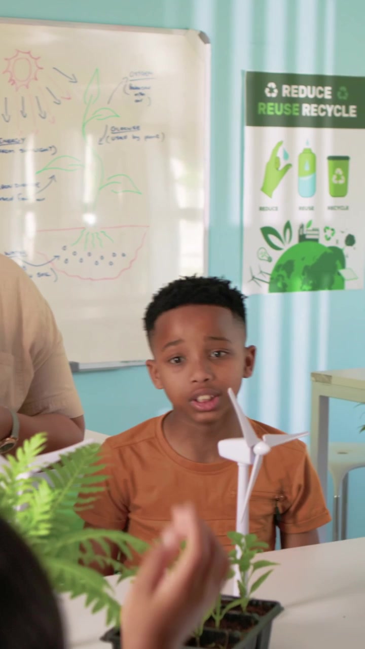 Vertical video: Teacher with tablet urging orange shirt boy checking eco turbine with lens in class