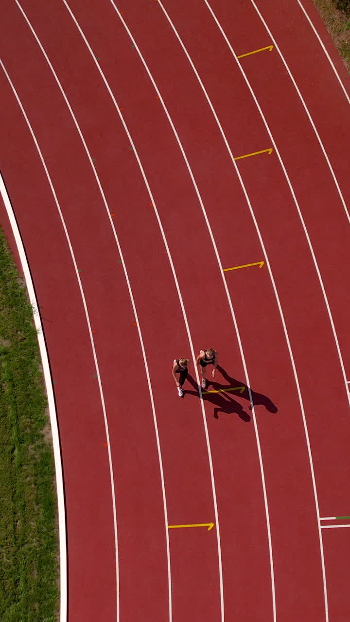 High angle vertical shot of two women jogging together on a professional red running track at a stadium during a sunny day. Ideal for fitness and sports content