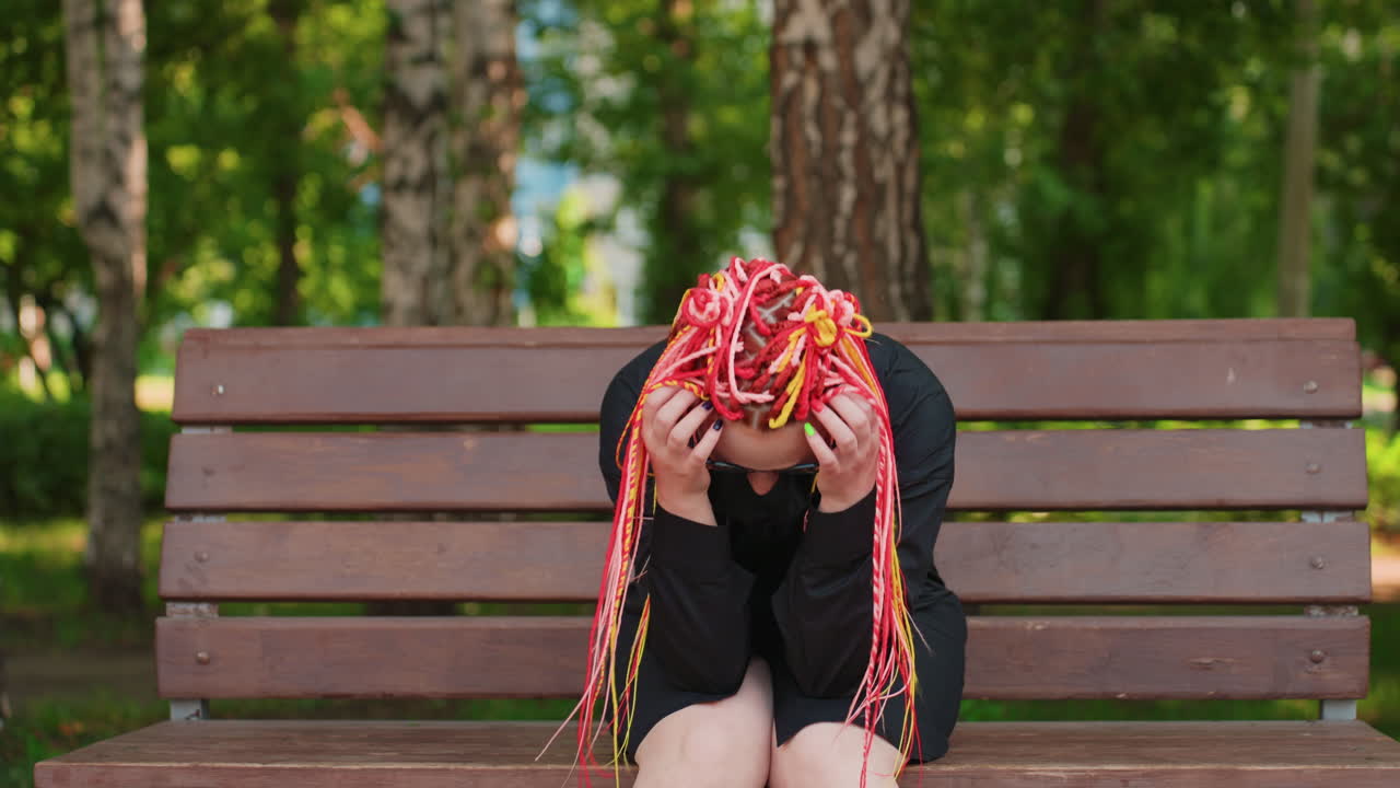 네온 브레이드를 한 백인 여성이 나무 벤치에 쓰러져 머리를 숙이고 있고, 다채로운 긴 브레이드 머리가 얼굴을 가리고 있으며, 배경에는 자작나무가 있고, 늦여름 햇살이 비치는 가운데 슬픔과 고립감을 전달하는 솔직한 인물 사진