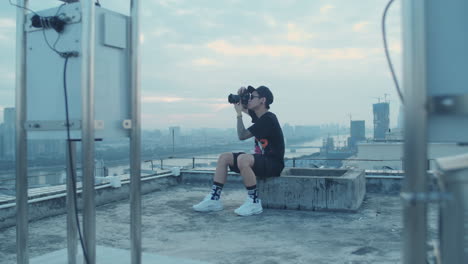 Offene-Zeitlupenaufnahme-Eines-Jungen-Asiatischen-Fotografen,-Der-Die-Innenstadt-Der-Stadt-Einfängt,-Während-Er-Am-Frühen-Morgen-Auf-Dem-Dach-Sitzt