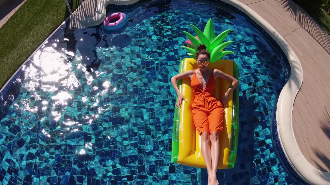 Young woman wearing orange jumpsuit and sunglasses relaxing on yellow inflatable pineapple shaped mattress in blue swimming pool during summer vacation, aerial view from above