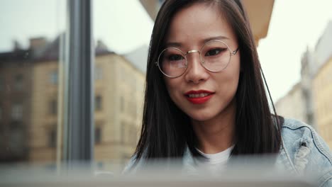 노트북 컴퓨터의 카페 테라스에 있는 테이블에 앉아 안경을 쓴 젊고 예쁘고 웃는 소녀가 화면을 보고 웃고 있습니다.
