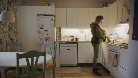 mother carrying her baby on a sling moves around kitchen cooking, wide shot