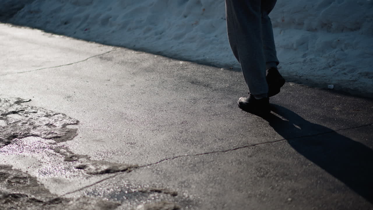 leg level view shows people in boots walking along tiled pavement beside snowbank, winter sun casts long shadows across wet asphalt, crisp cold air, urban sidewalk perspective, moody afternoon light