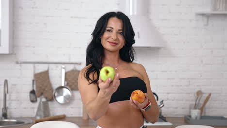 attractive female proposes fresh apples indoors. portrait of a woman with bare shoulders and belly holding juicy fruit and showing on camera in the kitchen.