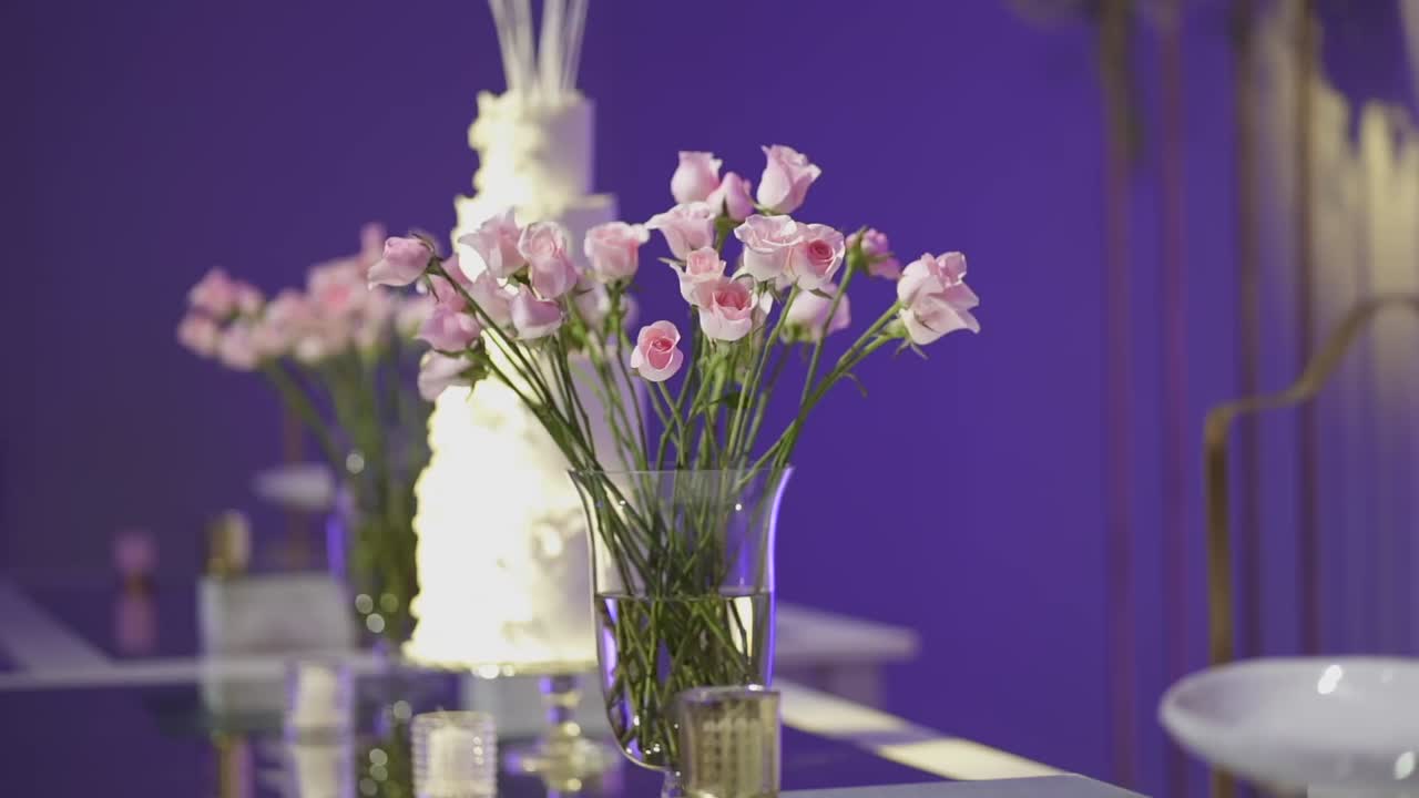 Camera tracking left showing a white wedding cake decorated with pink roses in vases, set in a room with purple ambient lighting and a blurred background