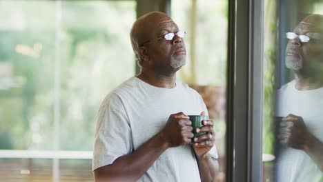 Thoughtful-african-american-senior-man-standing-drinking-mug-of-coffee-and-looking-out-of-window