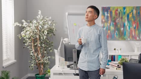 a man is standing in middle of company office holding cup of coffee, worker in a shirt is thoughtful, he is looking around the room, something is bothering him, he is stressed about work