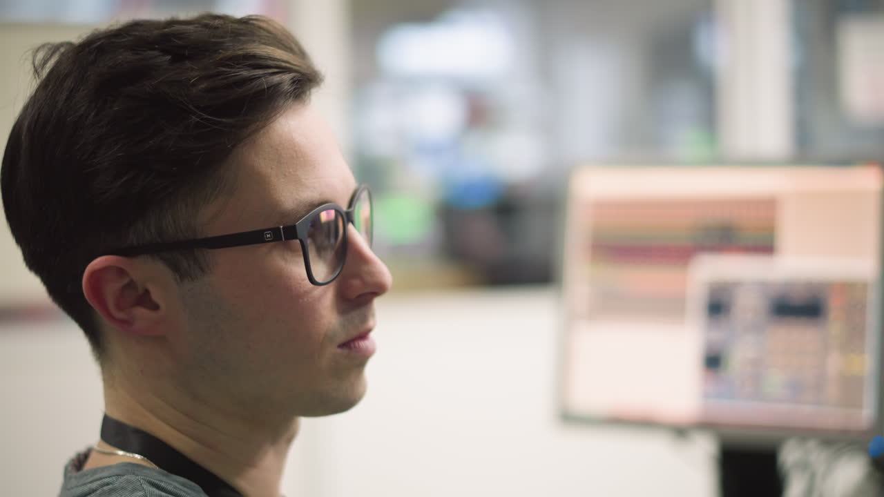Focused sound engineer adjusting audio levels at control desk with faders and knobs for a live broadcast, with audio equipment and multiple screens in the background. Professional audio work environment