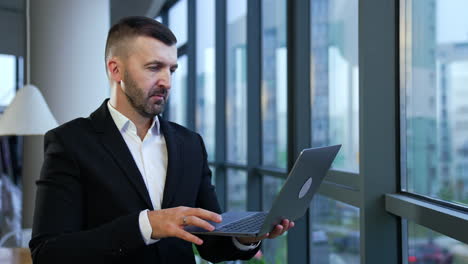 Caucasian man wearing black suit stands in the office and holds the laptop. Man with a personal computer in front of the panoramic window at work.