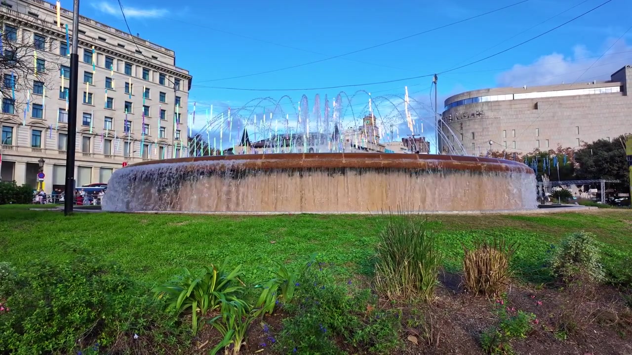 Fuente circular monumental en la Plaza de Cataluña, Barcelona, Cataluña, España, con chorros de agua en cascada rodeada de jardines y edificios emblemáticos en el centro de la ciudad, paneo lateral lento
