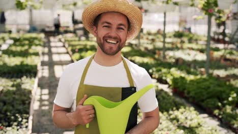 Portrait-of-a-young-male-gardener-in-apron-and-hat-showing-thumb-up-holding-watering-can-in-greenhouse