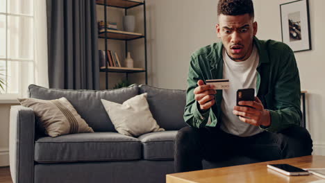 Young Man Looking Concerned While Using Credit Card and Smartphone for Online Transaction at Home