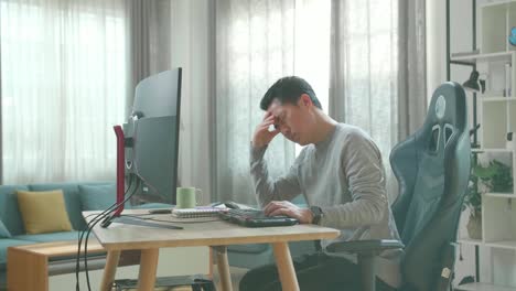 asian man in long sleeved t-shirt and black pants being tired while using the desktop computer for working at home.