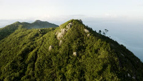 Drone-Distanciando-Un-Pico-De-Montaña-De-Bosque-Tropical-En-Un-Hermoso-Día-De-Verano-Revelando-El-Paisaje-Con-Océano-Por-Todas-Partes