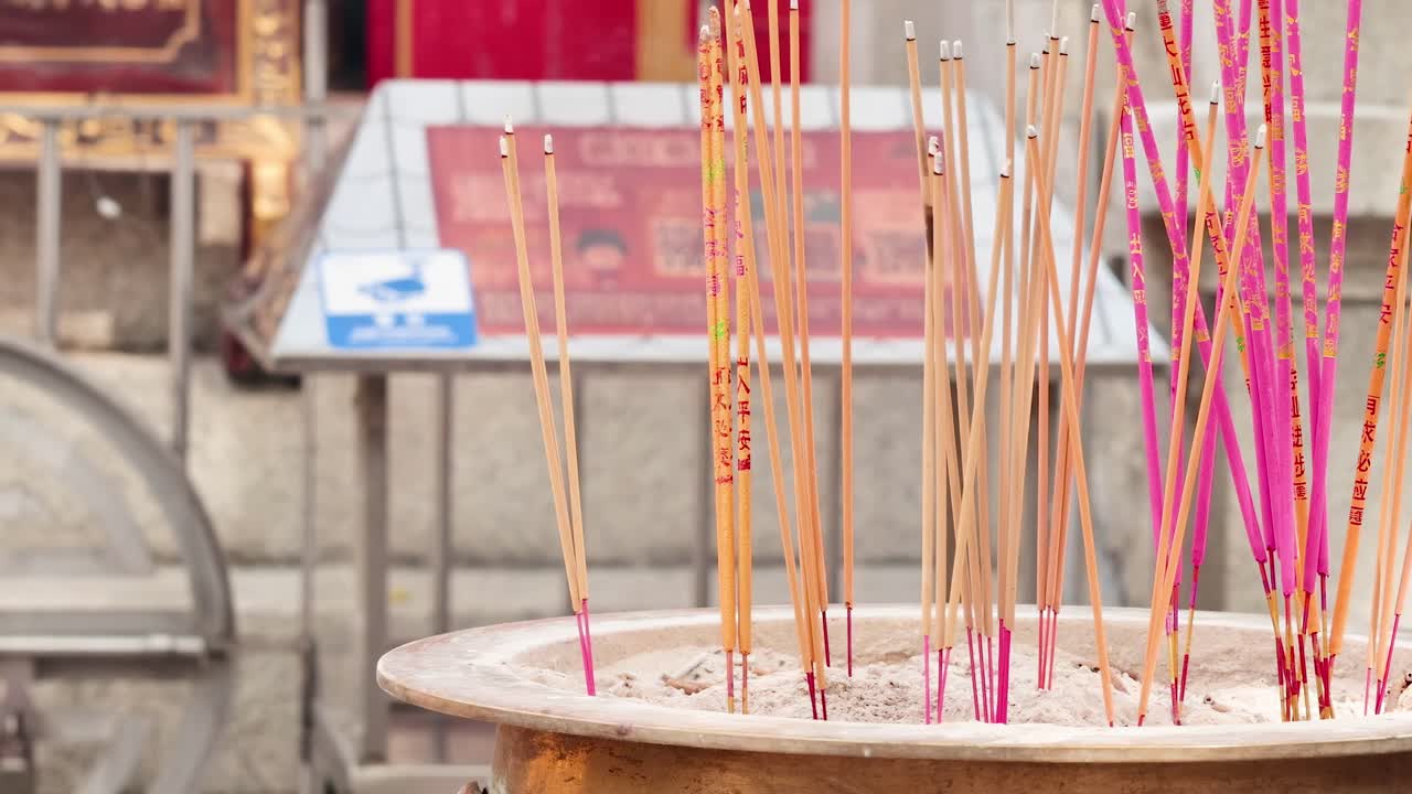 Colorful incense sticks stand upright in a circular holder, set against a temple backdrop.