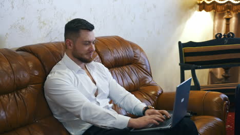 handsome businessman with a laptop in hotel. young bearded man is sitting on a comfortable sofa next to suitcase and working on a laptop at business trip.