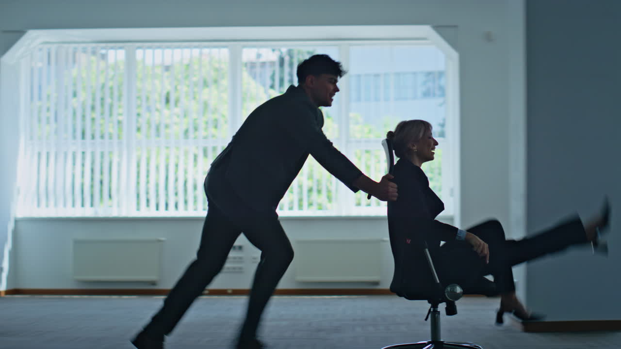Man colleague riding woman on office chair sharing laughter in corporate office