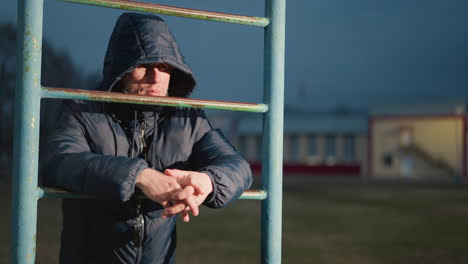 close-up of a person in a hooded jacket resting on an iron bar with clasped hands, illuminated by light from the background, looking focused