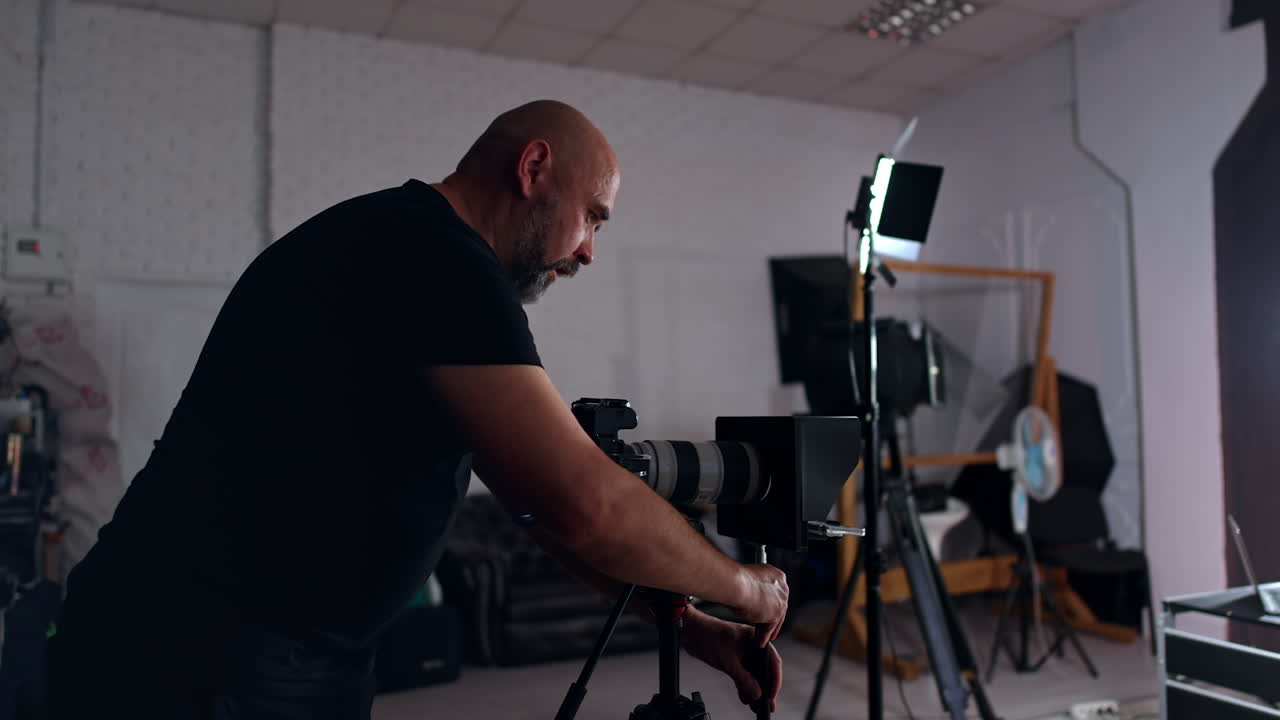 Bearded cameraman stands at the camera on tripod preparing it for work. Getting ready for footage in studio.