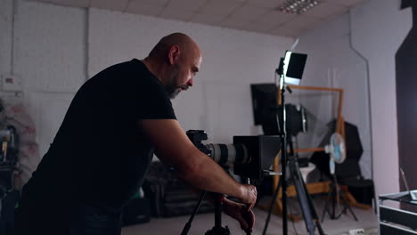 bearded cameraman stands at the camera on tripod preparing it for work. getting ready for footage in studio.