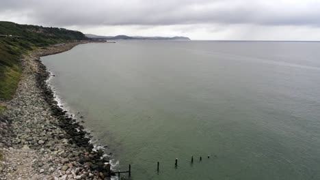 Lush-rocky-North-Wales-hillside-shoreline-ocean-boulders-aerial-view-slow-right-dolly-over-ocean