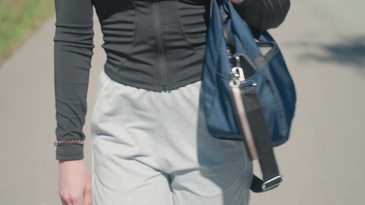 Primer plano de mujer caminando al aire libre mientras sostiene un bolso azul. La mujer viste ropa deportiva casual y lleva un bolso de mano en su mano derecha, con una suave iluminación natural que ilumina la escena.