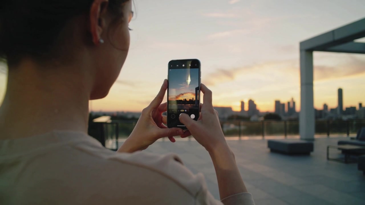 Mulher fotografando com o celular em um terraço na cobertura ao pôr do sol, ajustando o enquadramento e capturando a paisagem urbana.
