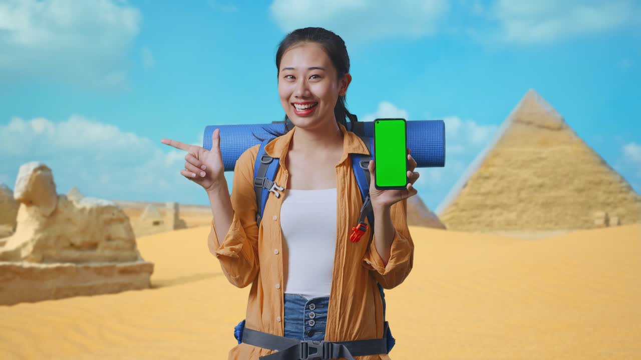 Asian Female Hiker With Mountaineering Backpack Showing Mock Up Green Screen Smartphone And Pointing To Side While Traveling In Pyramid Of Giza