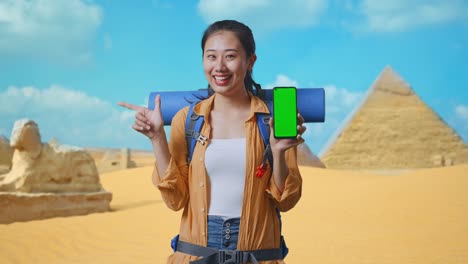 asian female hiker with mountaineering backpack showing mock up green screen smartphone and pointing to side while traveling in pyramid of giza