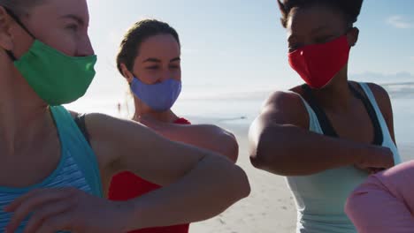 grupo de diversas amigas que usan máscaras faciales y se tocan los codos en la playa