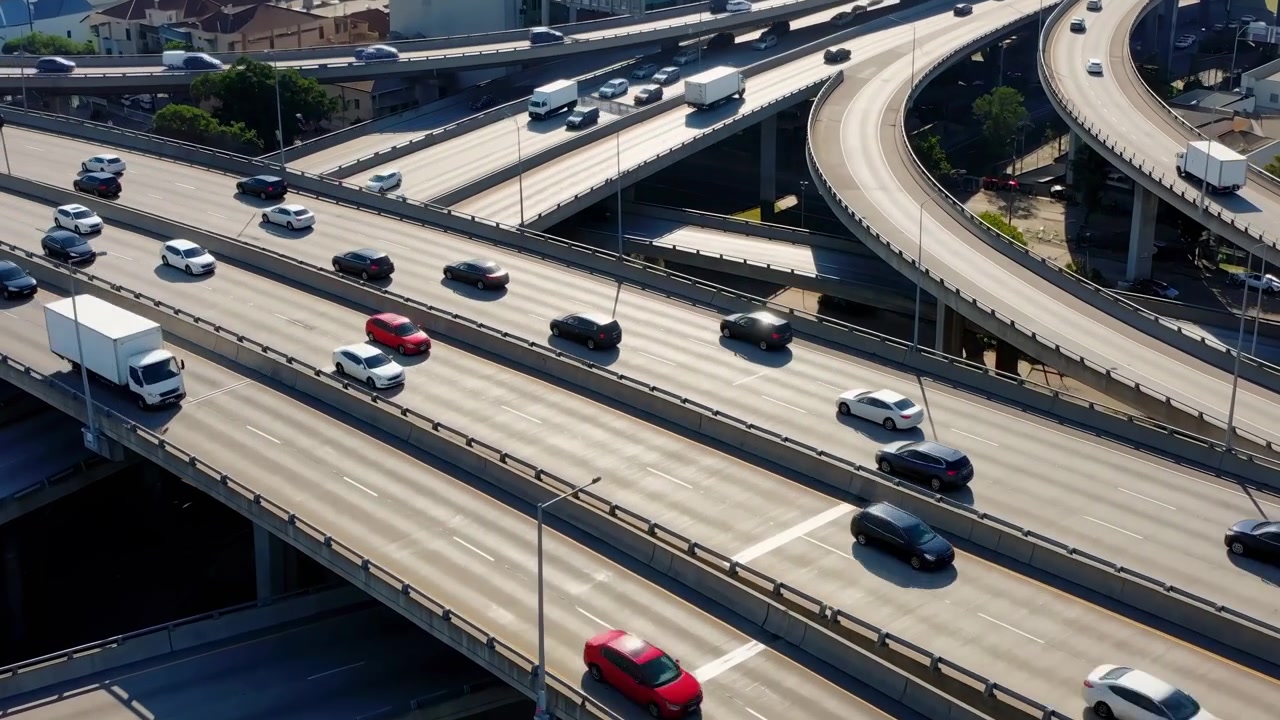 Una vista dall'alto mostra traffico costante che si muove in direzioni opposte su livelli autostradali sovrapposti, mentre i veicoli seguono le segnaletiche di corsia, le barriere di cemento e le rampe ampie accanto agli edifici urbani