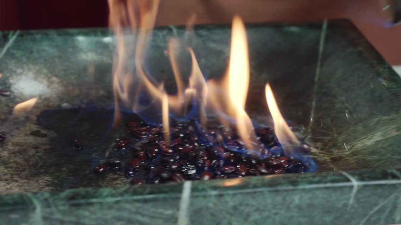 Närbild på en baristas hand som kastar en kopp kaffebönor med brinnande örtlikör ner i en grön stenplatta. Dramatiska flambéflammor flammar upp under den hantverksmässiga tillredningen.