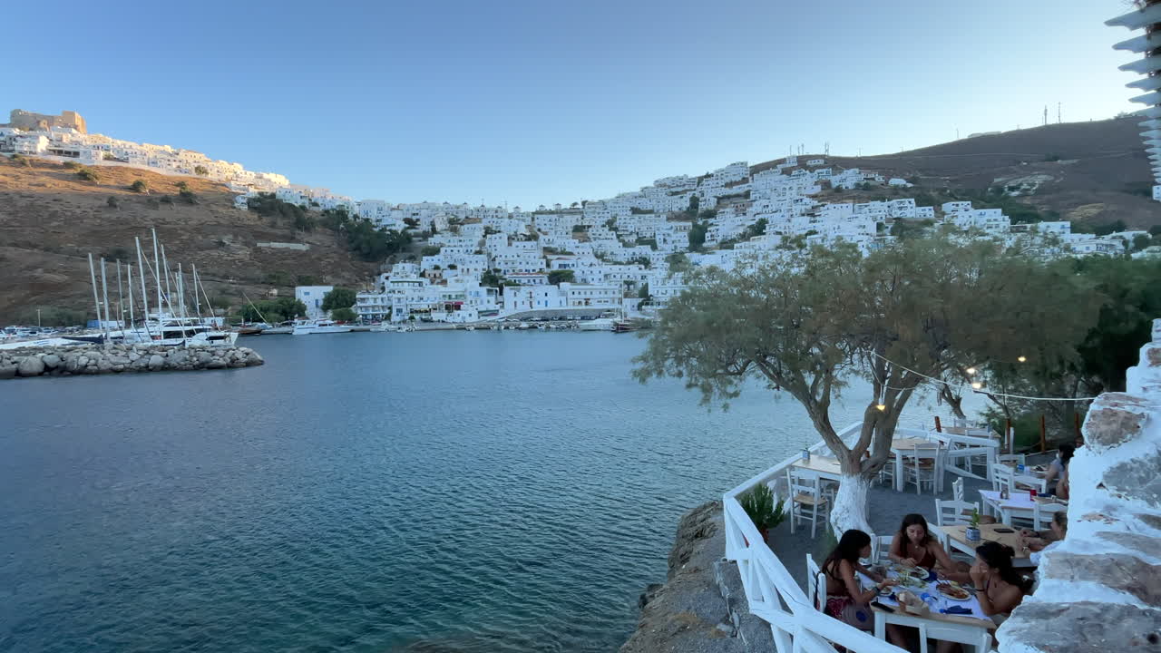 Greece, Astypalea Island, people having dinner at a restaurant with the view of the harbor, town and the medieval castle at the hill during sunset. Beautiful view on a quite late afternoon.