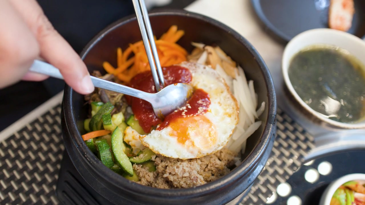 Seseorang menggunakan sendok dan sumpit untuk mencampur bibimbap dalam mangkuk batu panas.