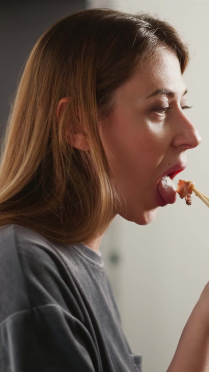 Primo piano di una donna che usa le bacchette per mangiare. La scena mostra la texture e il movimento delle sue mani che tengono utensili di legno, con un'illuminazione soffusa che evidenzia i dettagli nei capelli e nell'abbigliamento. Sfondo.