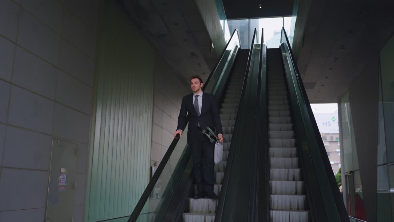Plano em câmera lenta com efeito paralaxe de um empresário profissional de terno escuro carregando um skate e uma bagagem enquanto desce uma escada rolante em um prédio de vidro moderno durante seu trajeto diário na cidade
