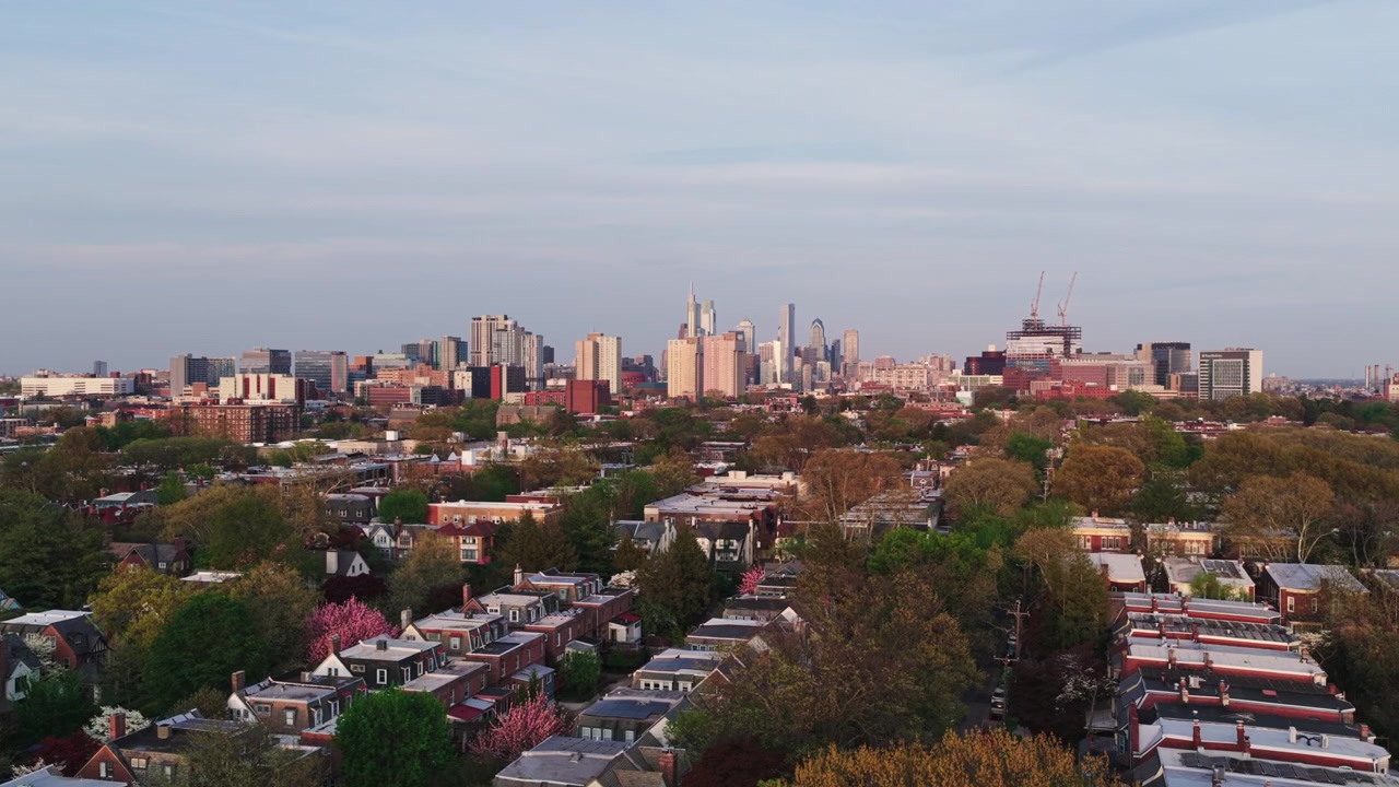 Vista aerea con drone di un quartiere di case a schiera di Filadelfia al crepuscolo, con fiori di ciliegio, la chioma degli alberi e l'intero skyline di Center City che brilla in lontananza.