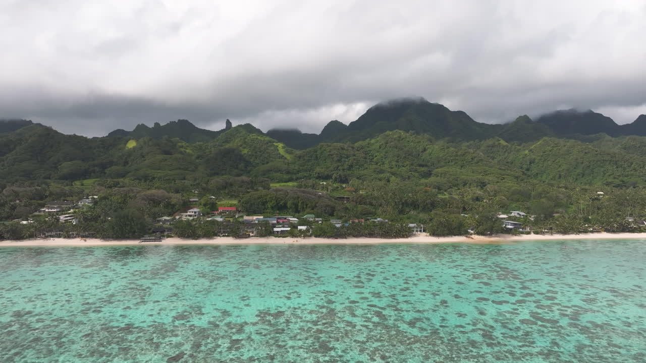 Aerial tracking left above coastline and homes built up below mountains in Cook Islands Rarotonga