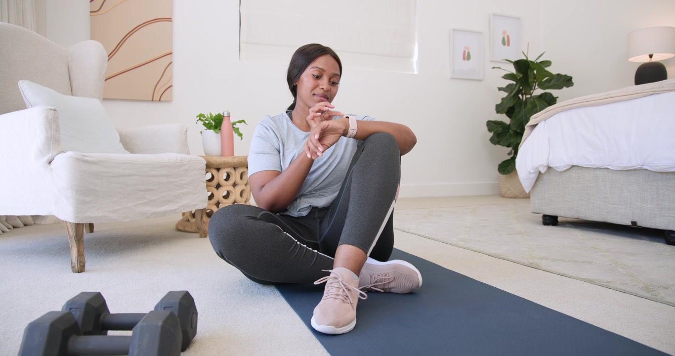 Donna africana che allaccia le scarpe da ginnastica su un tappetino blu a casa, poi controlla lo smartwatch per iniziare l'allenamento