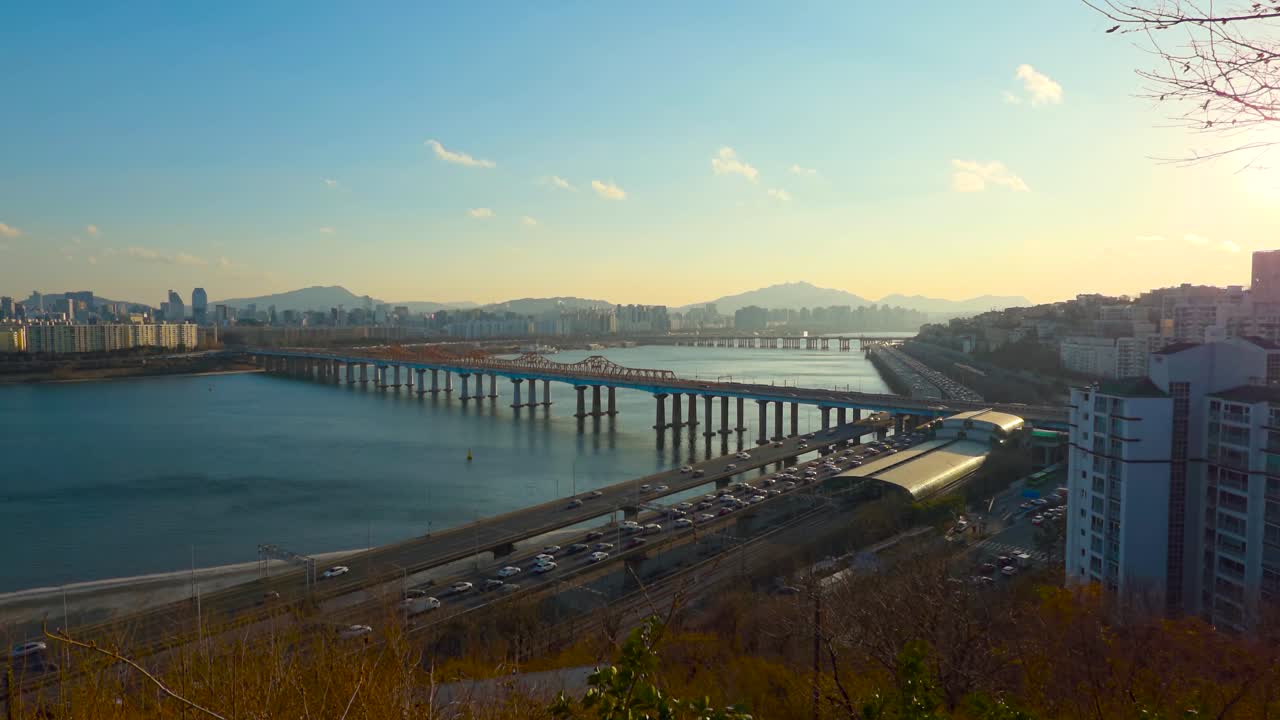 Zoom al tramonto in tempo reale da una collina che rivela il ponte Donho, la superstrada congestionata Gangbyeonbuk-ro e il tetto luminoso della stazione di Oksu lungo il fiume Han a Seul contro un sole al tramonto brillante